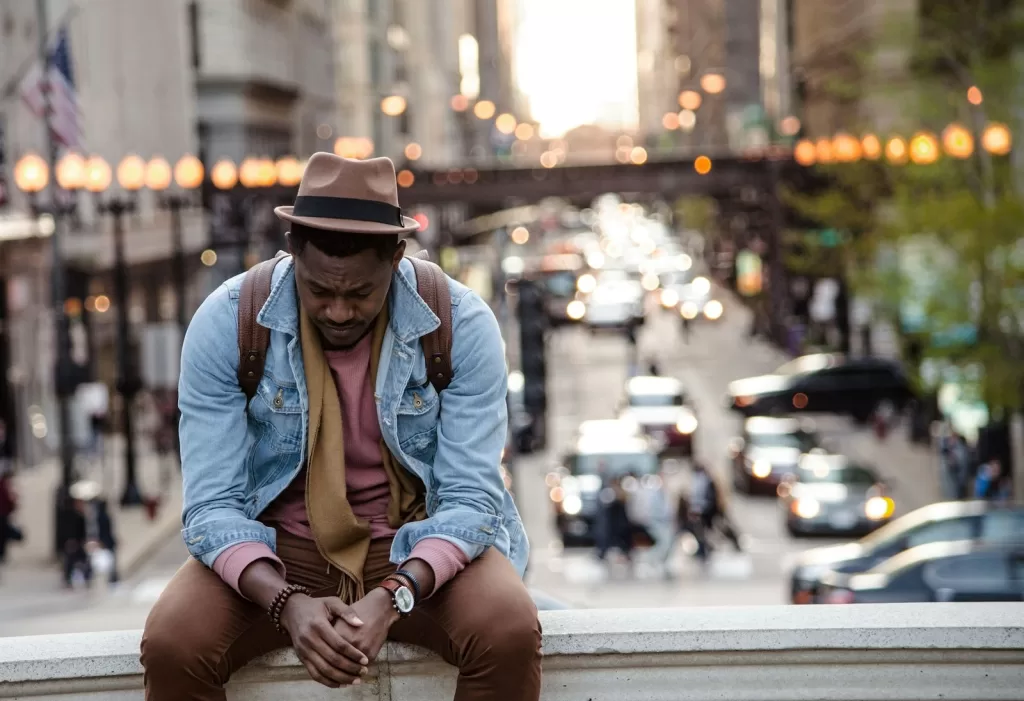 A man in a leather jacket looking down while sitting on a ledge in a city. Shows bad mental health